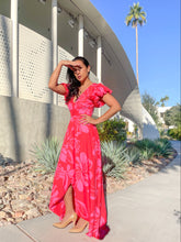 Woman in a red floral dress standing outdoors with a modern building and palm tree in the background.
