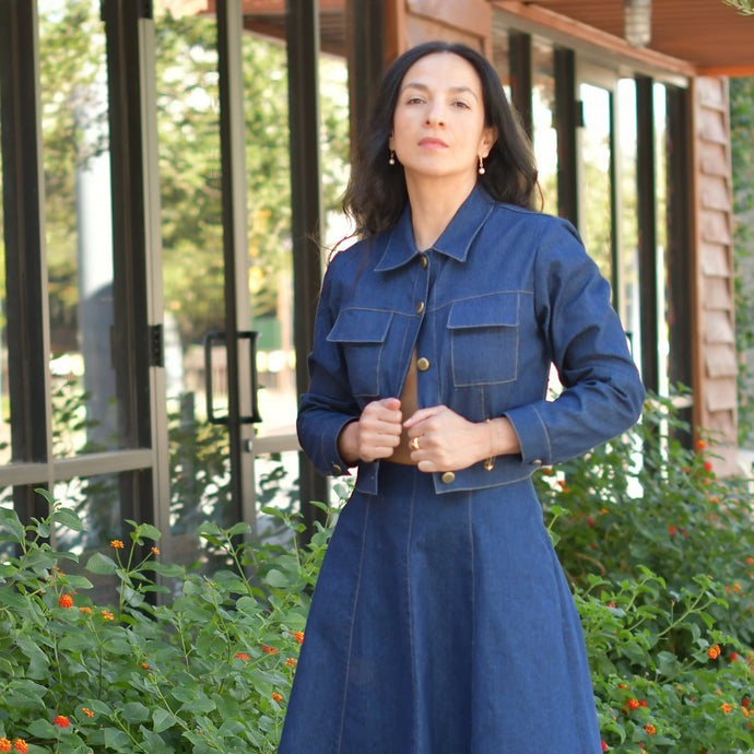 Woman in a blue dress standing against a wall with greenery and flowers.