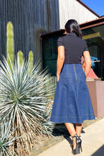 Woman walking outdoors wearing a black top and blue denim skirt, with a building and cactus in the background.